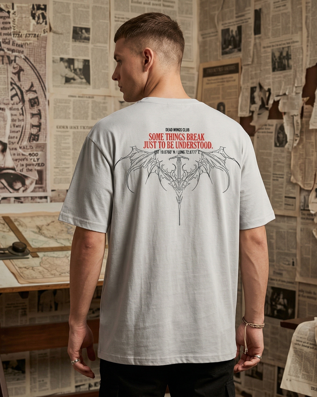 Man wearing a white t-shirt with text and graphics, standing in a room with newspaper walls and a globe.